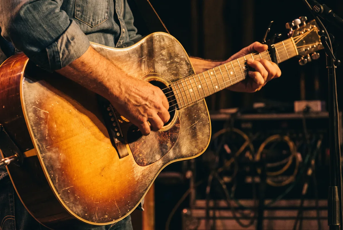 Close-up of a musician's hands on a worn sunburst acoustic guitar with a capo on the neck, lit by warm stage light