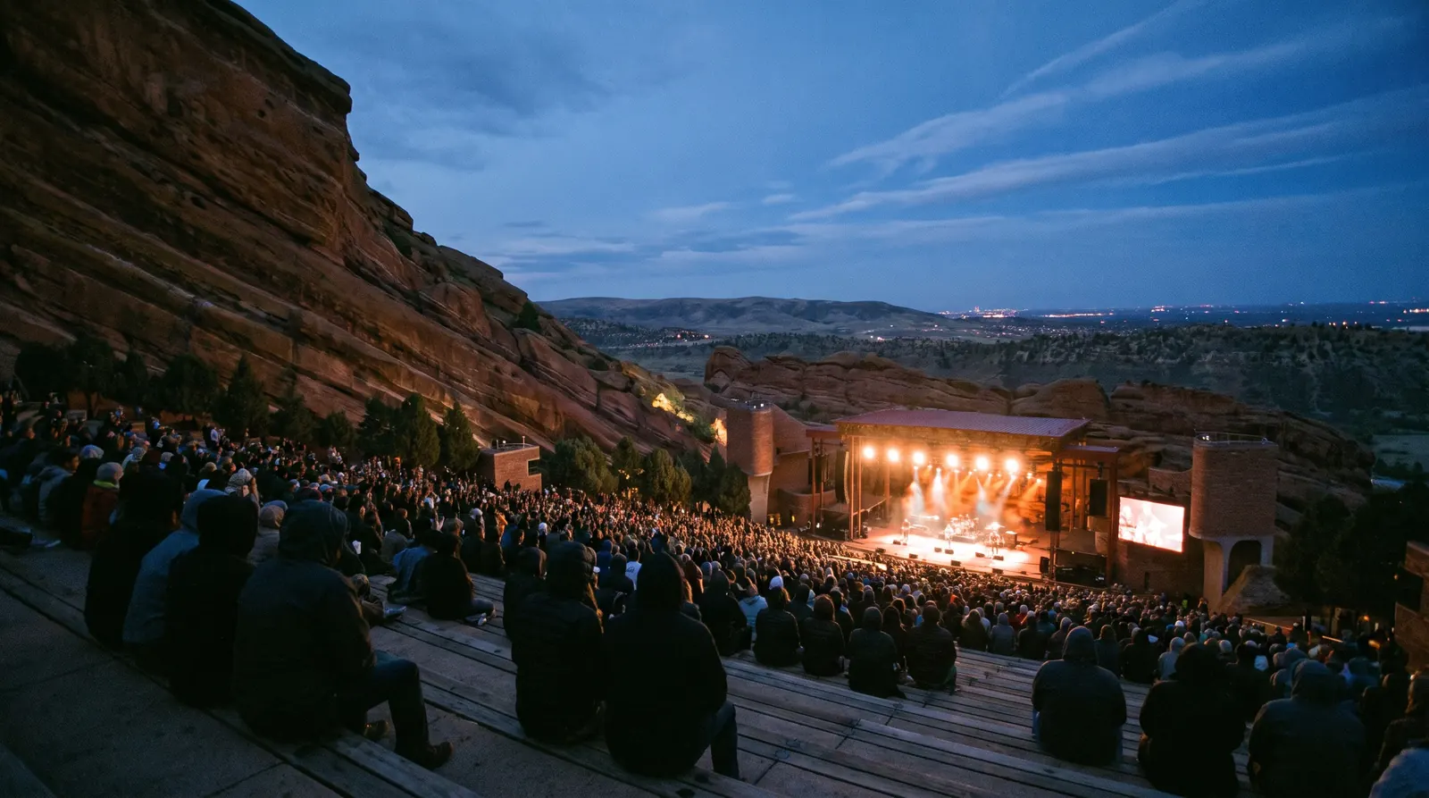 Tiered bench seating of a red sandstone amphitheater at dusk, full crowd facing a brightly lit stage between rock formations
